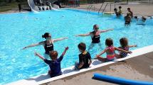 Two instructors in the pool demonstrating a skill to a group of children sitting along the edge of the pool.